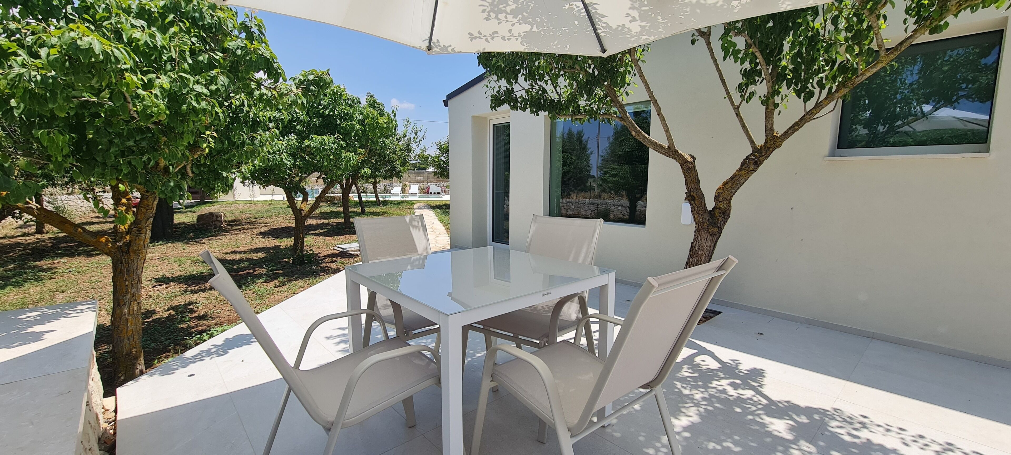 Eine Terrasse im Freien mit Glastisch und -stühlen, umgeben von Bäumen und einem modernen Gebäude.