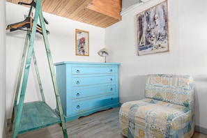 Um quarto com cômoda azul, cadeira estampada, escada e um quadro na parede.