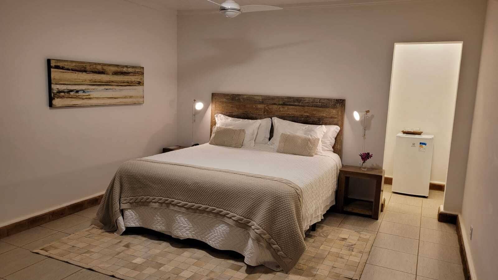 Quarto com cama, mesas de cabeceira, ventoinha no teto, obra de arte na parede e uma porta branca.