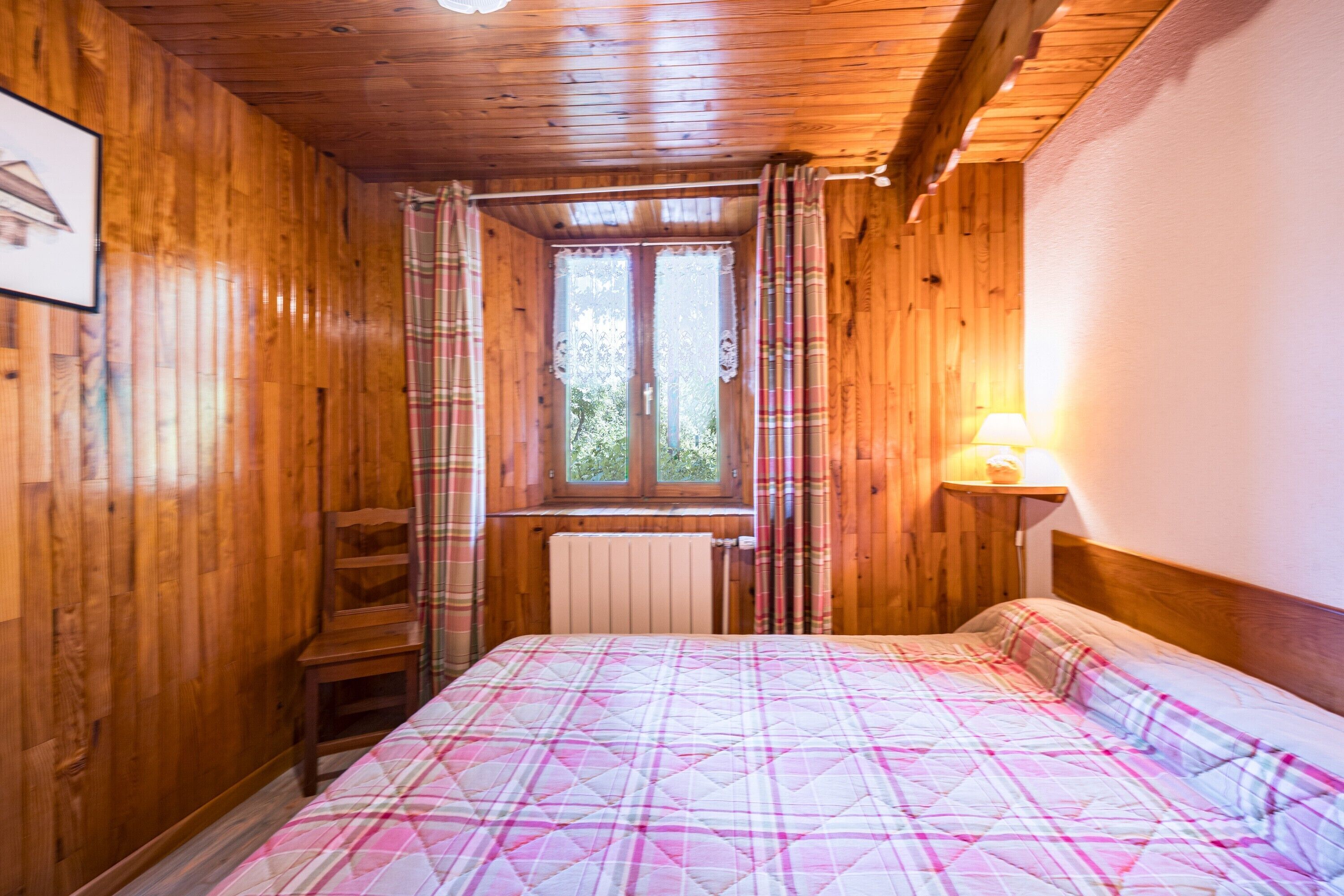 Ein Zimmer mit Holzboden, einem Bett, einem fenster mit Vorhängen, einem Heizkörper und einem Nachttisch mit Lampe.