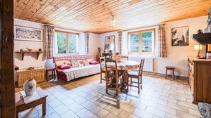 A cozy room with a wooden ceiling, a dining table with chairs, a sofa, a wicker side table, and a stone fireplace.