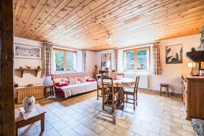 Une pièce chaleureuse avec un plafond en bois, une table à manger avec des chaises, un canapé, une petite table d’appoint en osier et une cheminée en pierre.