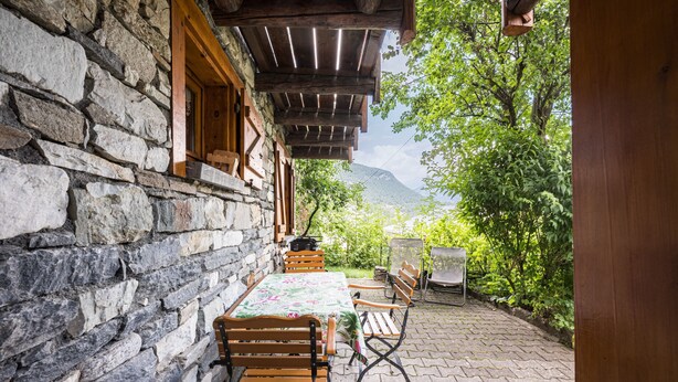 A stone building with a wooden table and chairs, a pergola, and a view of greenery and mountains.