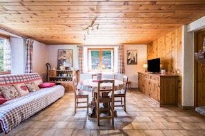 Interior acogedor de una cabaña con techo de madera, una mesa de comedor con sillas, un sofá, un televisor y una ventana con cortinas.