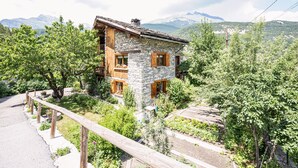 A stone building with wooden balconies, surrounded by greenery and mountains in the background.