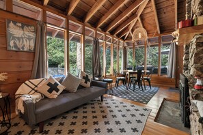 A cozy cabin interior with a wooden ceiling, large windows, a sofa with patterned cushions, a dining area, and a stone wall. - Clerestory Cottage (Big Bear Lake)