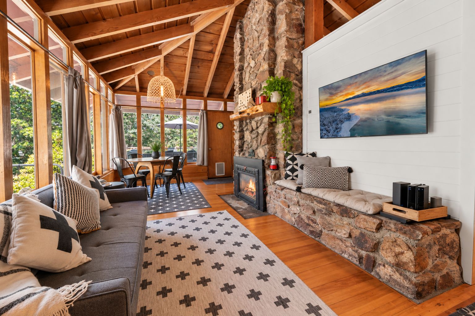 Un interior acogedor con chimenea de piedra, un conjunto de sofás, zona de comedor y un gran ventanal con vistas a la vegetación.