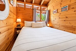 A wooden cabin room with a bed, nightstands, and a window with curtains. - Clerestory Cottage (Big Bear Lake)