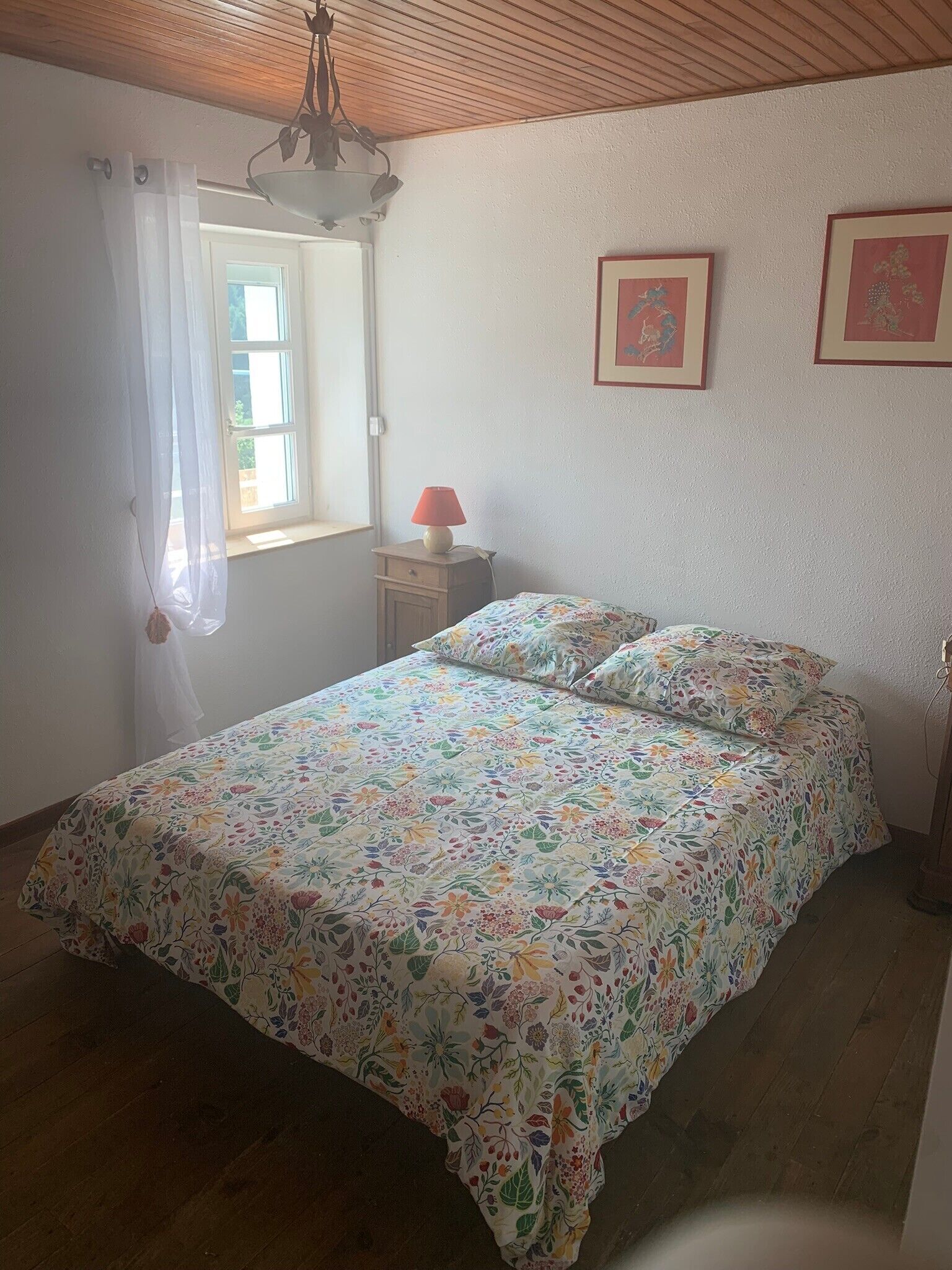 Um quarto com colcha estampada, teto de madeira, janela com cortinas brancas, abajur e duas molduras na parede.