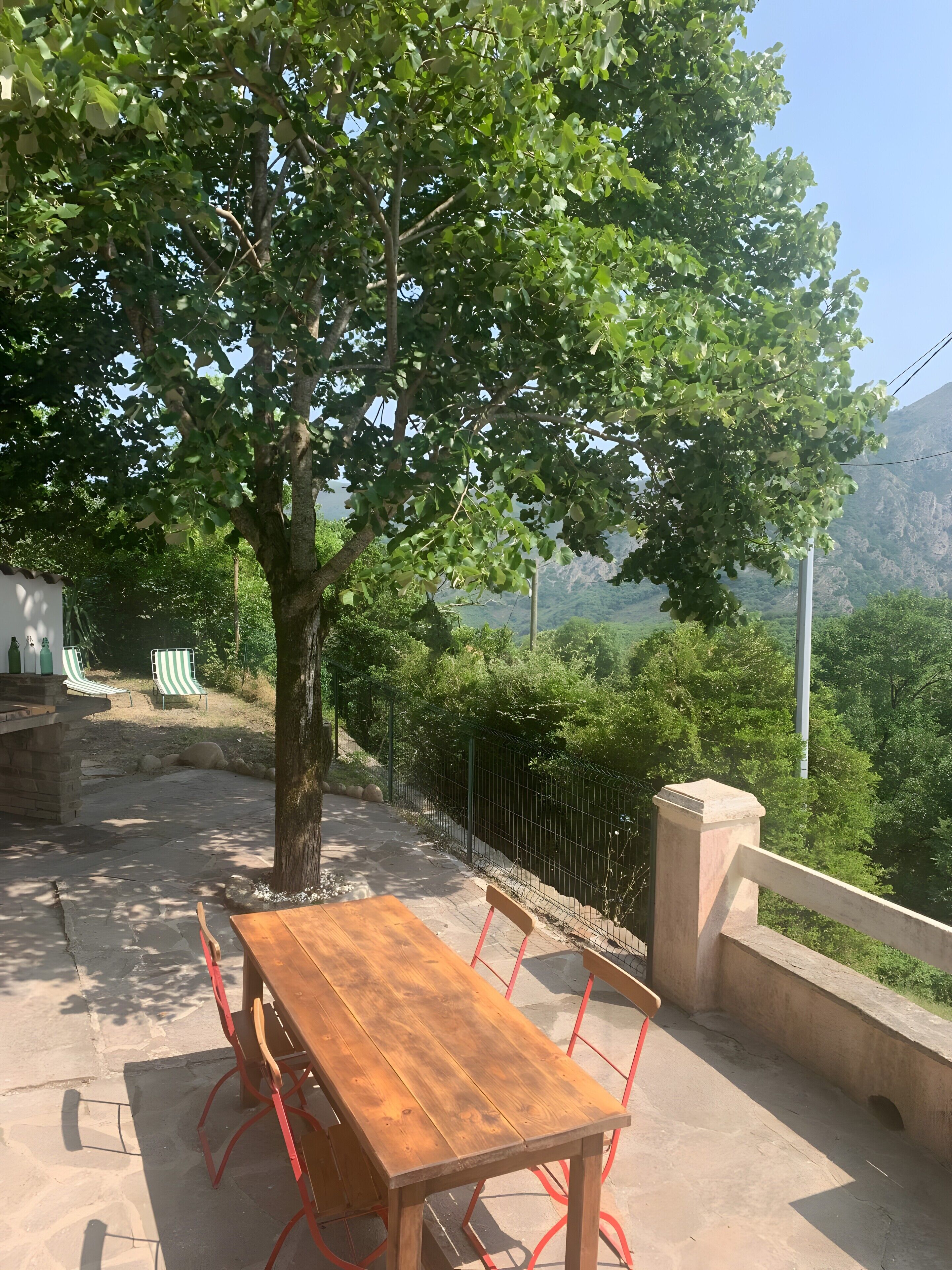 A wooden table with red metal chairs, a stone bench, and a view of a mountainous landscape.