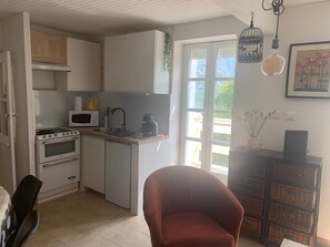 A compact kitchen with a stove, microwave, sink, and a small refrigerator. There is a wicker basket shelf and a chair in the room.