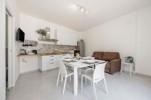 Une cuisine moderne avec des meubles blancs, une table à manger pour quatre personnes et un canapé marron dans l’espace salon.