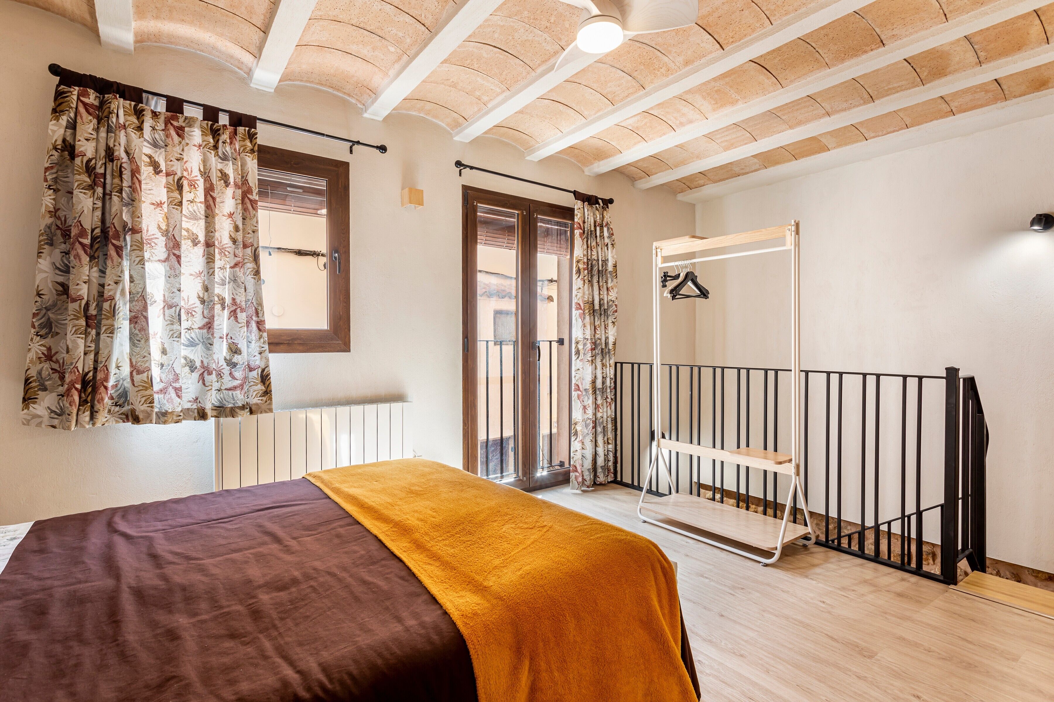 Un dormitorio con cama, ventana con cortinas, balcón con barandilla y puerta que da al exterior.