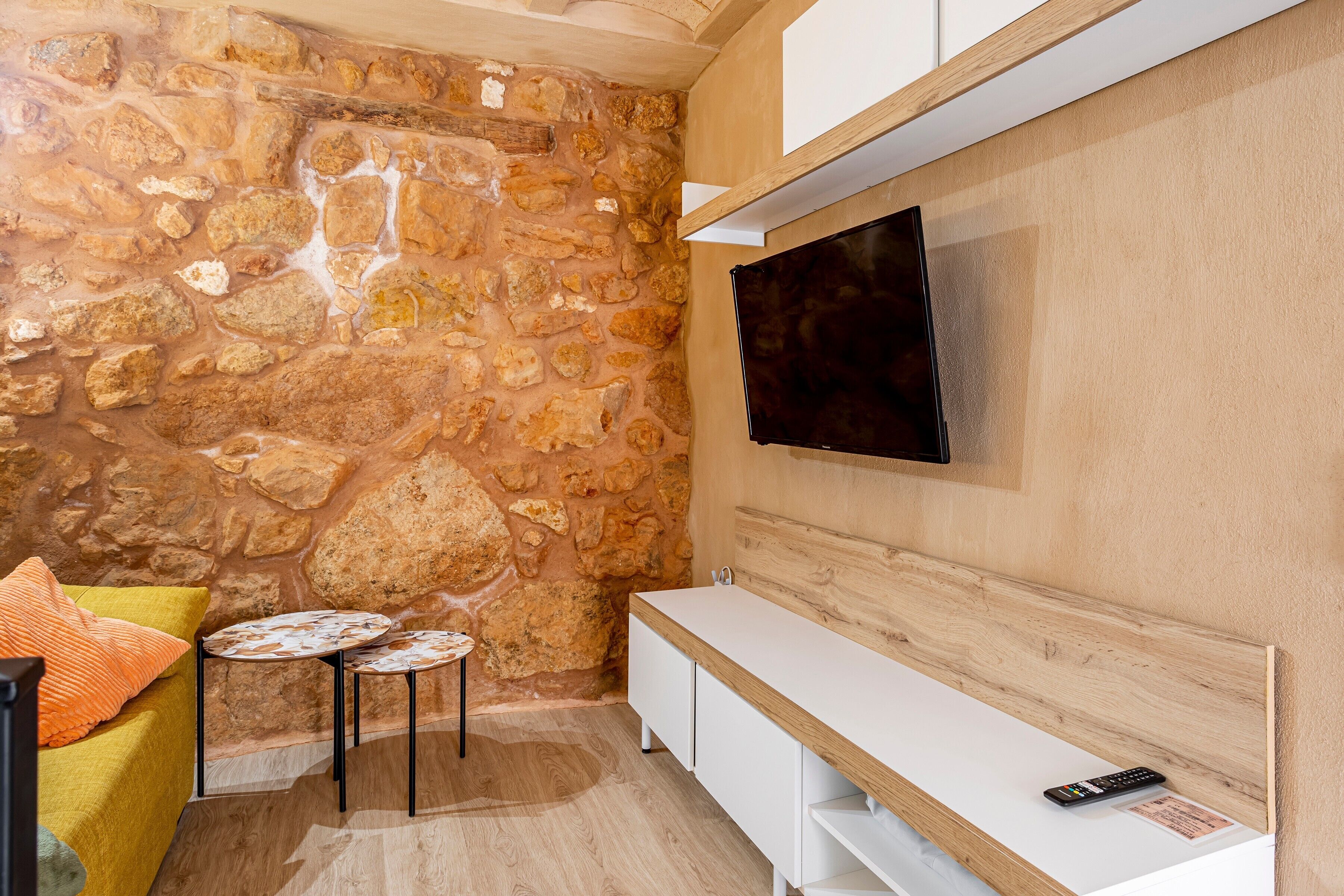 Ein Zimmer mit Steinwand, einem an der Wand montierten Flachbildfernseher, einer weißen Bank und einem kleinen runden Tisch mit Kissen.