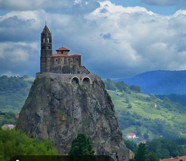 A historic stone tower with a bell, perched atop a rocky hill with a valley in the background.