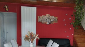 Un comedor con mesa de vidrio y sillas blancas, pared roja con una obra de arte de un árbol, y sofá negro.