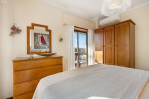 Quarto com cama, cómoda de madeira, pintura de um pássaro e vista através de uma porta de vidro.