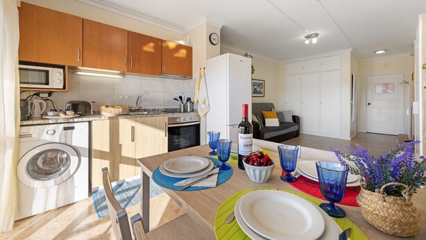 A compact kitchen with a washing machine, microwave, and toaster. There is a dining area with a table set for two, a bottle of wine, and a bowl of cherries.