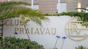 A sign with 'CLUBE PRAIAVU' and a logo, surrounded by greenery and a brick wall.