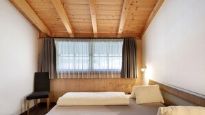 Une pièce avec des poutres apparentes en bois au plafond, un lit avec une literie blanche, une chaise et une fenêtre avec des rideaux.