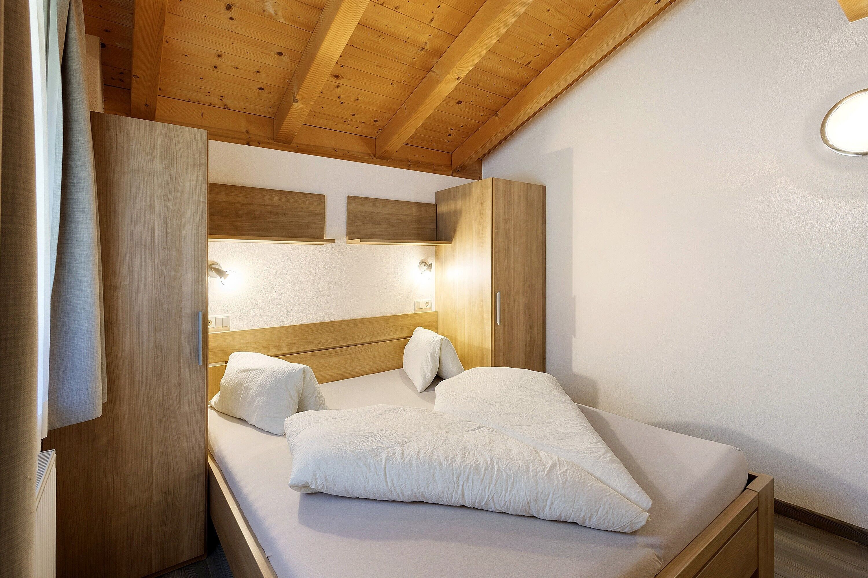 A bedroom with a wooden ceiling, a bed with white bedding, and a wardrobe.