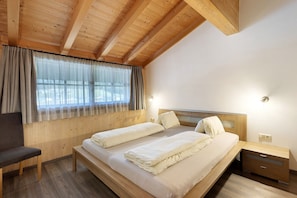 Une chambre avec un plafond en bois, un lit avec deux oreillers, une table de chevet, une chaise et une fenêtre avec des rideaux.