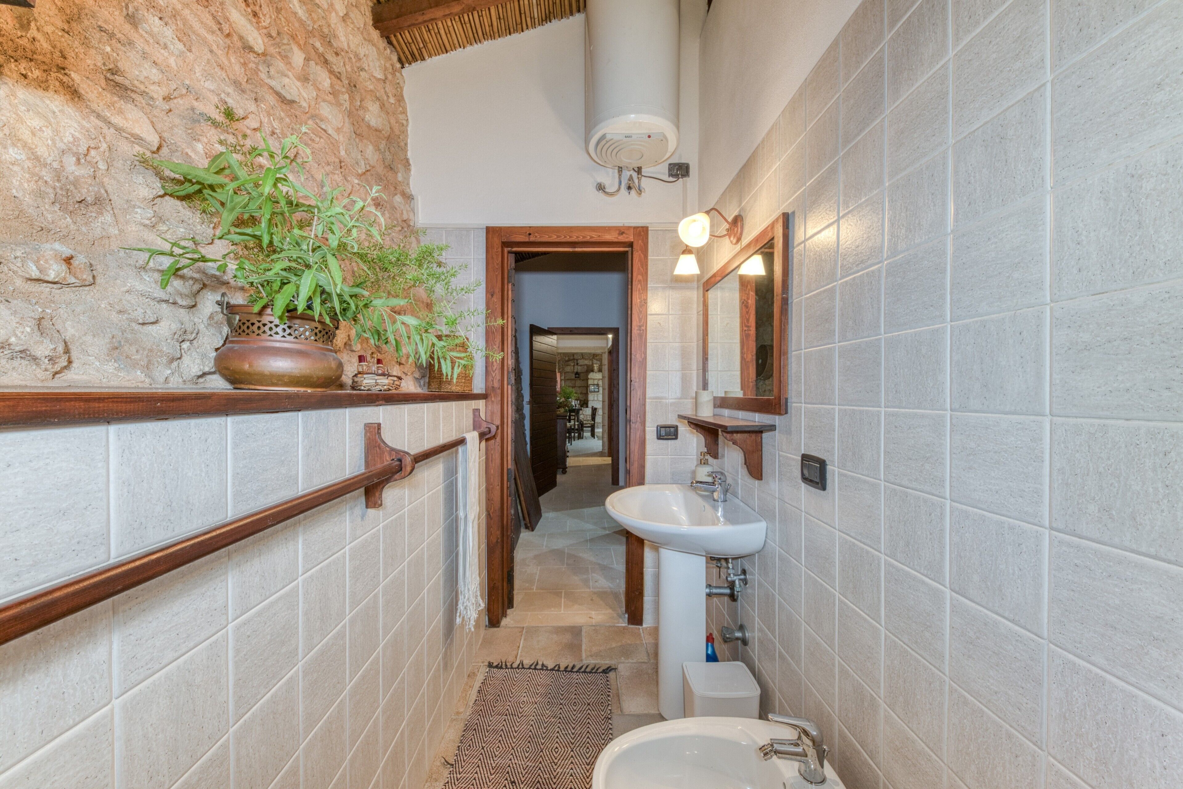 Un baño pequeño con pared de piedra, repisa de madera con plantas, lavamanos blanco y espejo.