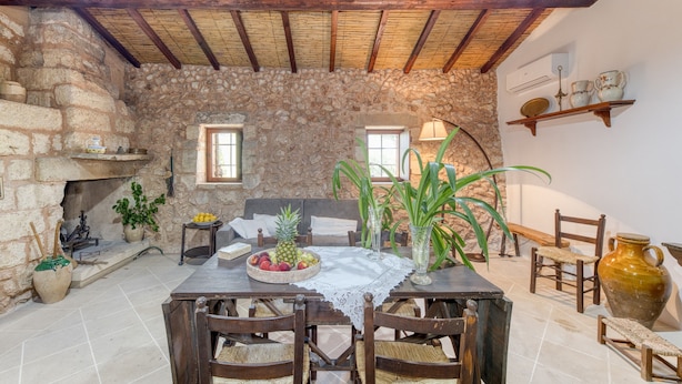 A rustic stone interior with a wooden table, chairs, a fireplace, and a stone wall.