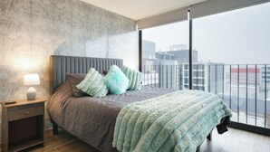 A modern bedroom with a large bed, a nightstand, a lamp, and a view of the city through large windows. - Modern and Cozy 1D1B Santiago Downtown Near Movistar Arena (Santiago)