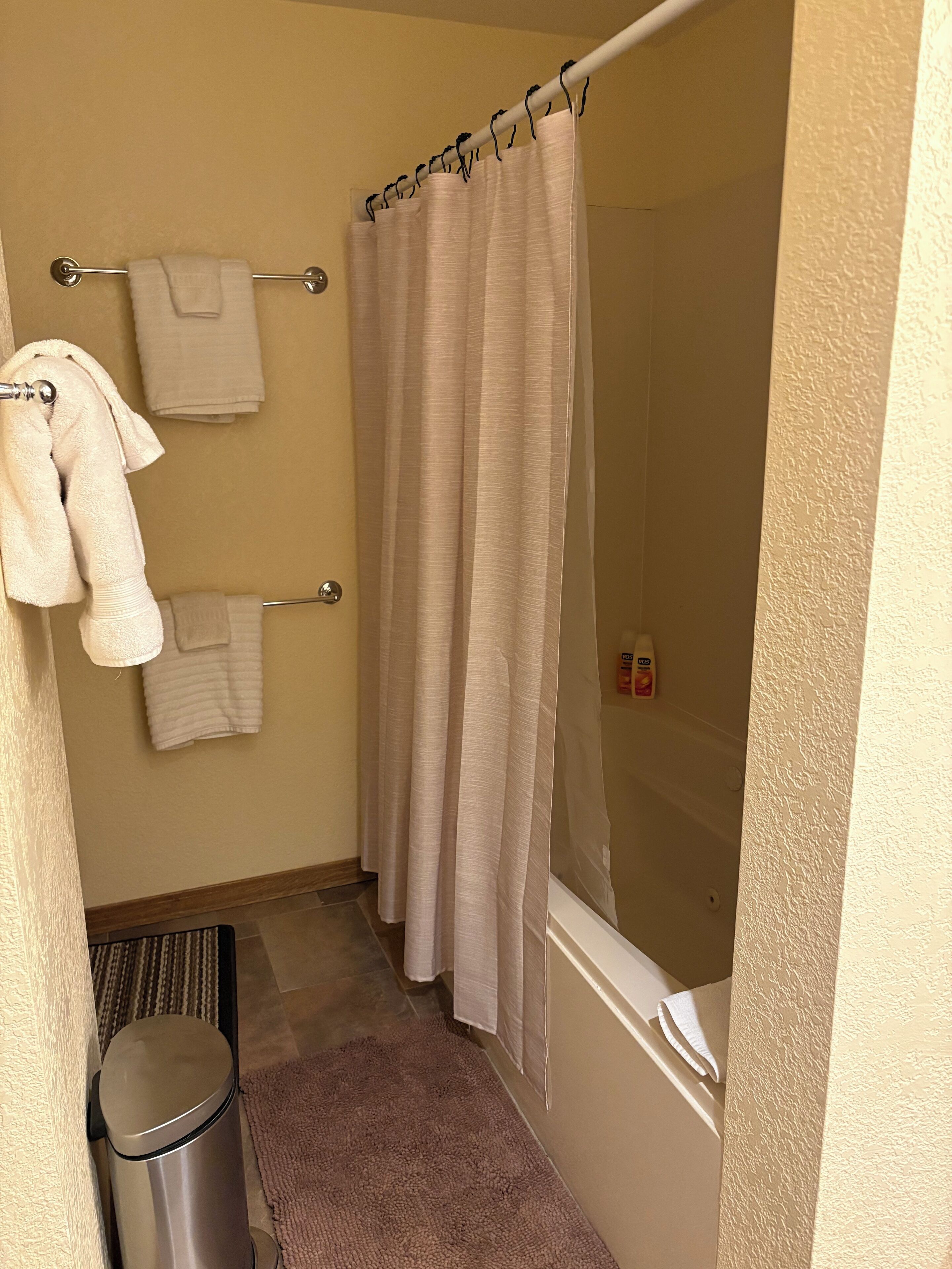 A bathroom with a shower curtain, towels on a rack, a trash can, and a window.