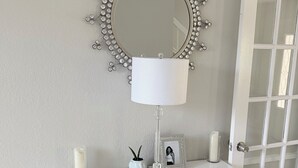 Een witte opbergkast met spiegel, een lamp, een ingelijste foto en decoratieve objecten op een dressoir.