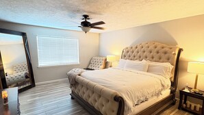 A bedroom with a tufted headboard, a wooden floor, a ceiling fan, a bedside table with a lamp, and a mirror on the wall. - Houston’s poolside vacation home (Houston)
