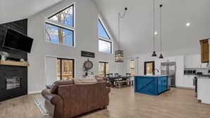 A spacious living area with a brown leather sofa, a fireplace, a dining table, and a kitchen with white cabinets and a blue island. - Lake House With Beach Access, Theater, Hot Tub (Port Austin)
