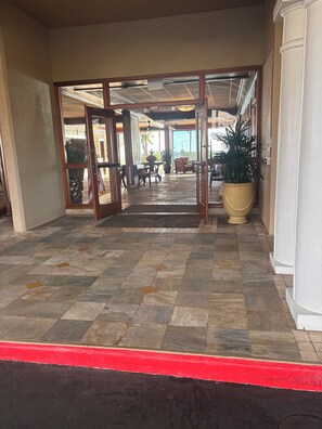 Ein Hotelzugang mit Fliesenboden, einer roten Bordsteinkante und Blick ins Innere, inklusive einer Sitzecke und einer großen Topfpflanze.