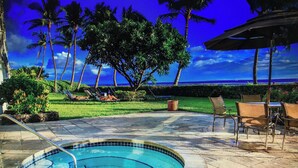Une piscine dont le bord est revêtu de carreaux, entourée de chaises longues et d’un espace patio offrant une vue sur l’océan et les palmiers.