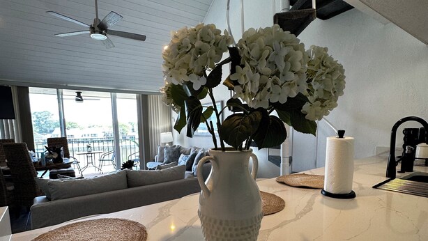 Un salon moderne avec un vase blanc rempli de fleurs posé sur un comptoir en marbre, un ventilateur de plafond et une vue sur l’extérieur grâce à de grandes fenêtres.