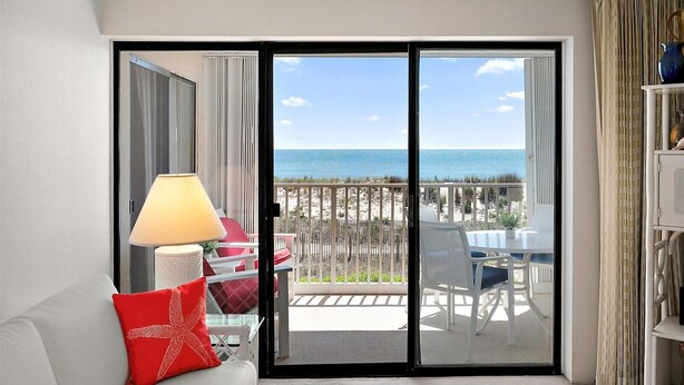 A room with a sliding glass door leading to a balcony with a view of the beach and ocean.