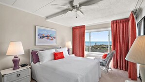 A hotel room with a bed, two bedside lamps, a ceiling fan, a window with red curtains, a beach-themed pillow, and a chair by the window.