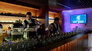 A bar with two servers attending to customers. The bar has a wooden interior with a modern design, including a slanted ceiling and a flat-screen TV mounted on the wall. - Hotel El-Arz (Boghni)