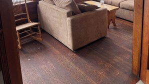 Ein Zimmer mit einer Couch, einem Schaukelstuhl und einem Holztisch. Große Fenster bieten Blick auf ein Gewässer.