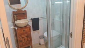 A bathroom with a wooden vanity, a round mirror, a beige sink, a toilet, and a glass-enclosed shower. - MINI HOUSE 25M2 (Barcelona)