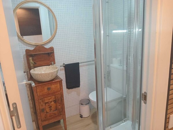 A bathroom with a wooden vanity, a round mirror, a beige sink, a toilet, and a glass-enclosed shower. - MINI HOUSE 25M2 (Barcelona)