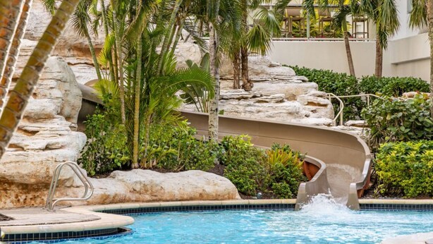 Una piscina con tobogán, rodeada de palmeras y rocas.