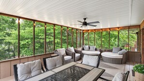 Ein überdachter Außenbereich mit Korbmöbeln, einem Deckenventilator und großen Fenstern mit Blick auf Bäume.