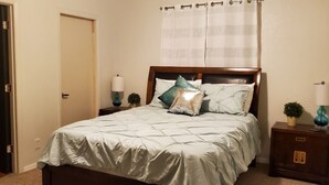 Un dormitorio con una cama, dos mesitas de noche, una mesita auxiliar, una puerta y una cortina.