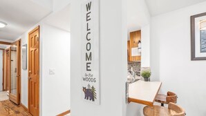 Un passadís amb un cartell que diu 'WELCOME to the WOODS', portes de fusta i una cuina amb taulell i tamborets.