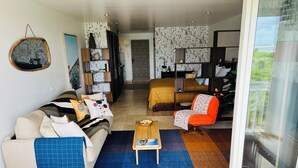 Un salon moderne avec un canapé, un fauteuil et une table basse. Une porte donne accès à une autre pièce, et une porte vitrée offre une vue sur l’extérieur.
