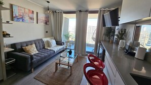 A modern living room with a grey sofa, a glass coffee table, and red chairs. There is a TV mounted on the wall and a large window with curtains. - Apartamento Reñaca Live (Viña del Mar)