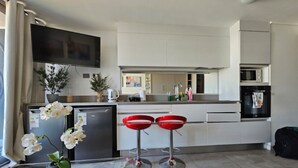 A modern kitchen with white cabinets, a stainless steel refrigerator, a microwave, and an oven. There are two red bar stools and a small table with a vase of white orchids. - Apartamento Reñaca Live (Viña del Mar)