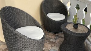 Dos sillas de mimbre con cojines blancos en un patio empedrado, una pequeña mesa redonda con una planta y una barandilla blanca.